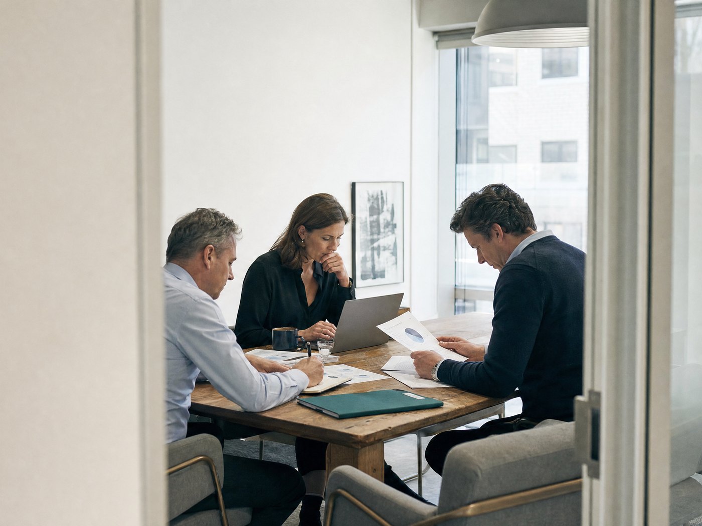 Three people at a wooden table in a glass meeting room, going through papers.